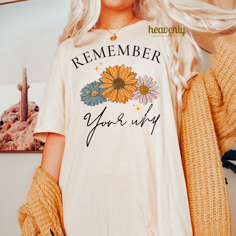 K&ouml;nnte beinhalten: Ein wei&szlig;es T-Shirt mit dem Text "REMEMBER your why" in schwarzer Schrift. Der Text ist von drei stilisierten Blumen in Blau-, Gelb- und Rosat&ouml;nen umgeben.