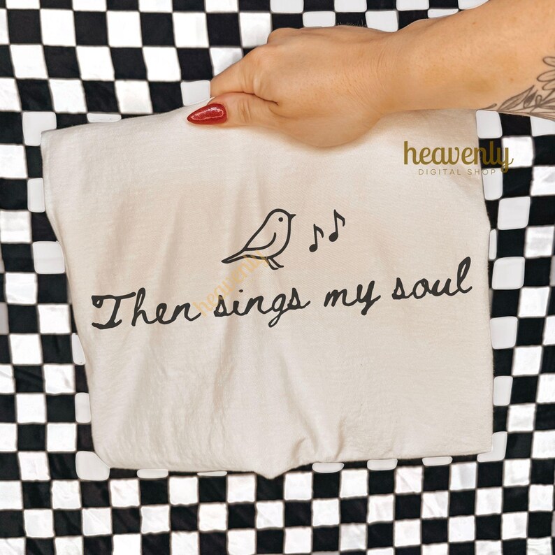 May include: A beige tote bag with the text "Then sings my soul" in black script. A black bird and musical notes are printed above the text. The bag is held up against a black and white checkered background.