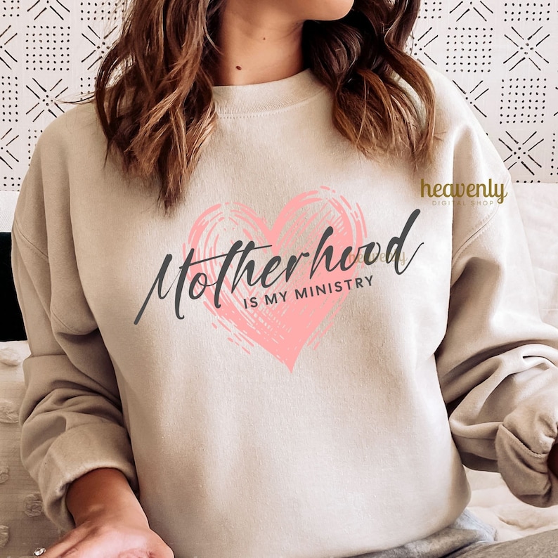 Op de afbeelding: Een beige sweatshirt met een roze hartontwerp en de tekst "Motherhood is my ministry".