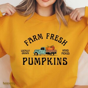 May include: Mustard yellow sweatshirt with a graphic design. The design features the words "FARM FRESH PUMPKINS" above a light blue truck carrying pumpkins. The text also includes "LOCALLY GROWN" and "HAND PICKED".