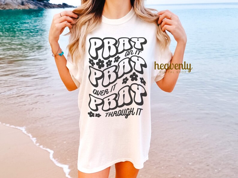 May include: Cream-colored t-shirt with the words "PRAY on it, PRAY over it, PRAY through it" in a black, bubble-style font. Small black flower graphics are interspersed throughout the text. The shirt has a relaxed fit.