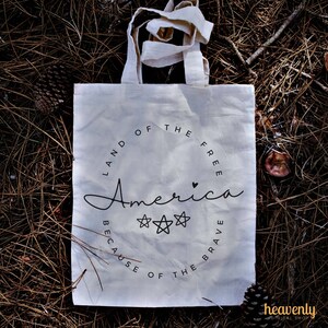 May include: White canvas tote bag with black text that reads "Land of the Free, America, Because of the Brave". The text is in a circle with three stars and a small heart.