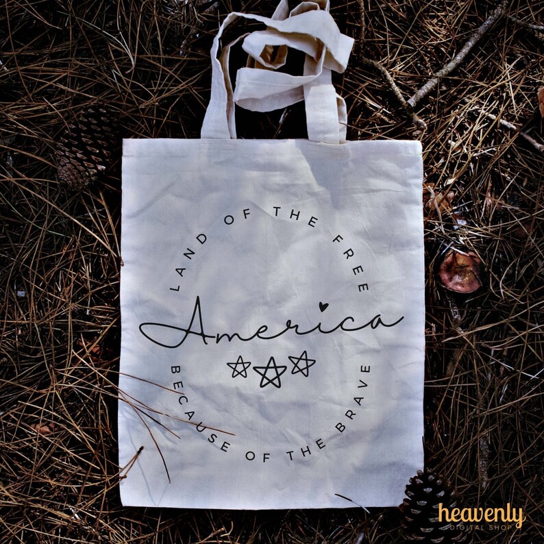 May include: White canvas tote bag with black text that reads "Land of the Free, America, Because of the Brave". The text is in a circle with three stars and a small heart.