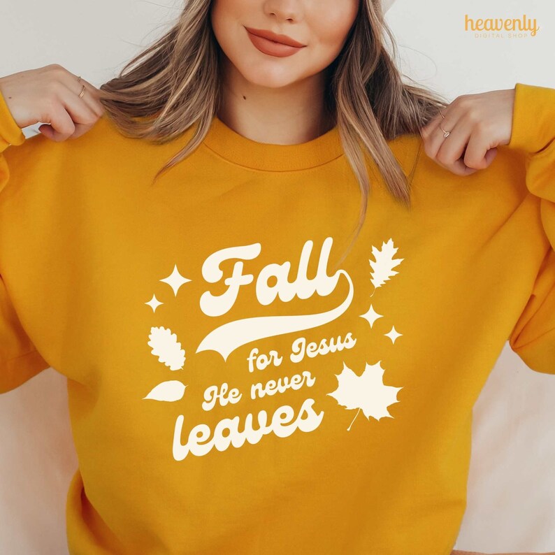 Op de afbeelding: Een geel sweatshirt met een witte afbeelding waarop staat "Fall for Jesus He never leaves" met gestileerde bladeren en sterren.