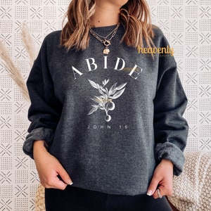 May include: A dark gray sweatshirt with the word "ABIDE" in white lettering, a stylized floral design, and the text "JOHN 15" in white lettering.