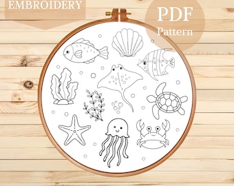 May include: A hand embroidery PDF pattern featuring various sea creatures, including fish, a stingray, a turtle, and a crab, all in a simple black and white line art style. The design is set within a wooden embroidery hoop.