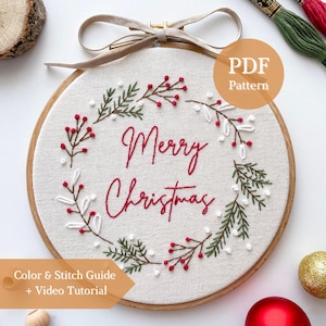 Puede incluir: Un bastidor de bordado circular de madera con tela color hueso y un diseño de corona bordado a mano. La corona presenta ramas de pino verdes, bayas blancas y bayas rojas que rodean el texto bordado en rojo Merry Christmas.
