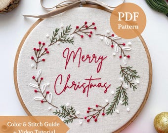 May include: A circular wooden embroidery hoop containing off-white fabric with a hand-stitched wreath design. The wreath features green pine branches, white berries, and red berries surrounding the red embroidered text Merry Christmas.
