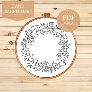May include: A circular wooden embroidery hoop containing white fabric with a black line art pattern of an oak leaf and acorn wreath. The text HAND EMBROIDERY and PDF TEMPLATE are visible in the corners of the image, framing the central hoop.