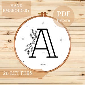 Pode incluir: Um bastidor de bordado de madeira com tecido branco apresentando a letra "A" preta com detalhes botânicos. Os textos "HAND EMBROIDERY", "PDF Pattern" e "26 LETTERS" também são visíveis.