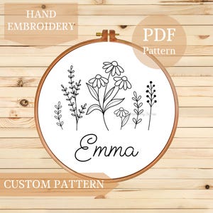 Könnte beinhalten: Ein runder Stickrahmen aus Holz mit weißem Stoff, der ein schwarzes Strichzeichnungsdesign verschiedener Wildblumen und den Namen Emma in Schreibschrift zeigt. Der Text HAND EMBROIDERY, PDF Pattern und CUSTOM PATTERN ist zu sehen.