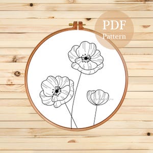May include: A black and white floral embroidery pattern in a wooden hoop. The design features three poppy flowers with detailed petals and stems. The text "PDF Pattern" is in a circle.