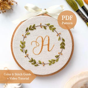 May include: An embroidery hoop containing light-colored fabric with a hand-stitched orange letter A in the center, surrounded by a circular wreath of green leaves and small orange berries. A cream-colored ribbon is tied at the top of the wooden hoop.