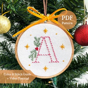 May include: A circular wooden embroidery hoop containing white fabric with a hand-stitched letter A in red and white candy cane stripes. The letter is accented with green pine sprigs, red berries, and gold starbursts. A bright yellow velvet ribbon is tied at the top.