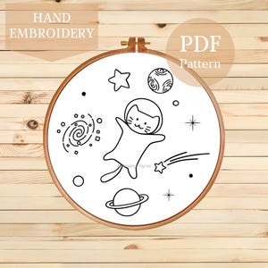 Könnte beinhalten: Ein Handstickrahmen mit einem schwarz-weißen Weltraum-Design. Das Design zeigt eine Katze als Astronaut, Planeten, Sterne und eine Galaxie. Der Text "HAND EMBROIDERY" und "PDF Pattern" sind ebenfalls enthalten.