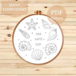 May include: A hand embroidery PDF pattern displayed in a wooden hoop. The design features a collection of seashells, a starfish, and the words "sea sand sun" in black. The text "Hand Embroidery" and "PDF Pattern" are also included in the design.