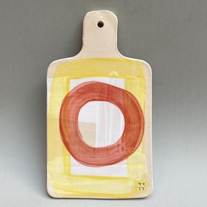 Könnte beinhalten: Ein gelbes und rotes Keramik-Schneidebrett mit einem Griff. Das Brett hat einen roten Kreis mit einem weißen Zentrum und ein gelbes Quadrat darum herum. Das Brett ist mit "NK 22" signiert.