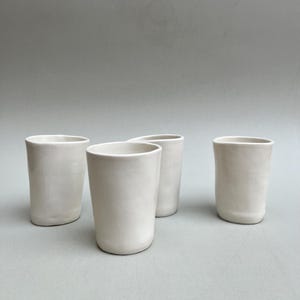 Könnte beinhalten: Vier weiße Keramikbecher mit leicht abgerundeter Form. Die Becher sind in einer Reihe auf einer hellgrauen Oberfläche angeordnet.