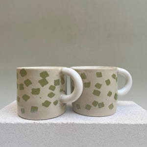 May include: Two ceramic mugs with white handles and a green geometric pattern. The mugs are sitting on a white surface.