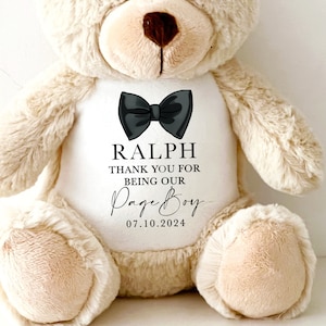 Op de afbeelding: Een beige teddybeer met een witte buik. De beer heeft een zwarte vlinderdas en de naam "RALPH" boven de tekst "THANK YOU FOR BEING OUR Page Boy 07.10.2024".