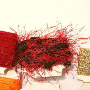 K&ouml;nnte beinhalten: Sortiment von Stickgarnrollen in Rot, Gold und Orange. Die rote Rolle ist ein flauschiger, strukturierter Faden. Die goldene Rolle ist ein gl&auml;nzender, metallischer Faden. Die orangefarbene Rolle ist ein glatter Baumwollfaden.