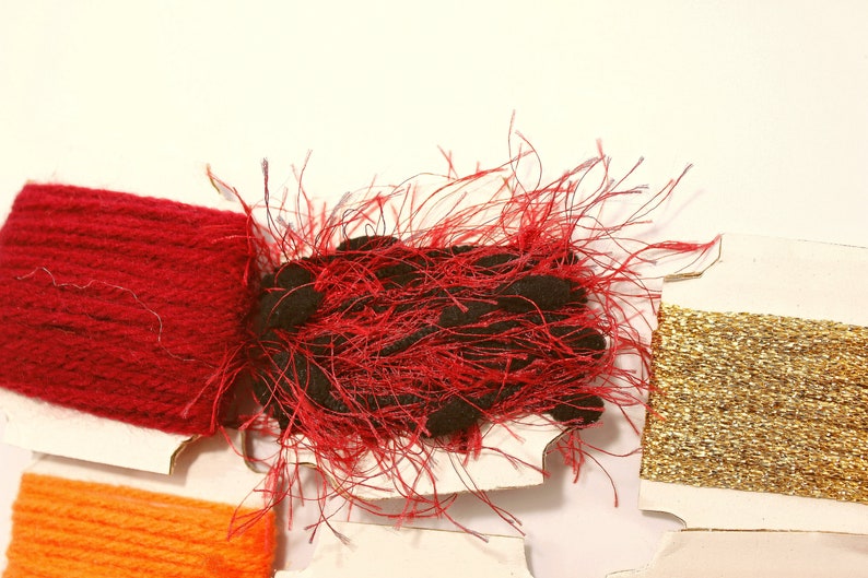 K&ouml;nnte beinhalten: Sortiment von Stickgarnrollen in Rot, Gold und Orange. Die rote Rolle ist ein flauschiger, strukturierter Faden. Die goldene Rolle ist ein gl&auml;nzender, metallischer Faden. Die orangefarbene Rolle ist ein glatter Baumwollfaden.