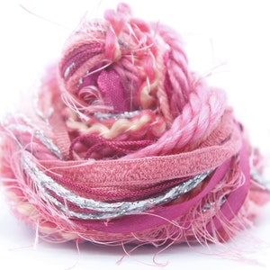 Peut inclure: Un peloton de laine rose et argenté avec une variété de textures et de fibres. La laine est enroulée de manière lâche et a un aspect duveteux.