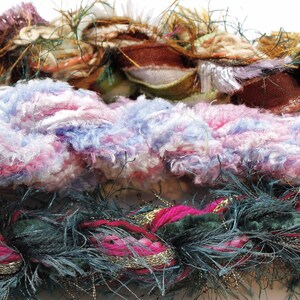 Può includere: Primo piano di tre tipi diversi di filato. Lo strato superiore è un filato marrone e beige con una trama scintillante. Lo strato intermedio è un filato rosa e blu con una trama soffice. Lo strato inferiore è un filato verde scuro e rosa con una trama soffice.