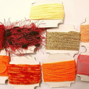 K&ouml;nnte beinhalten: Sortiment an bunten Garnrollen auf wei&szlig;em Karton. Die Garnfarben umfassen Rot, Orange, Gelb, Gold und Rosa.