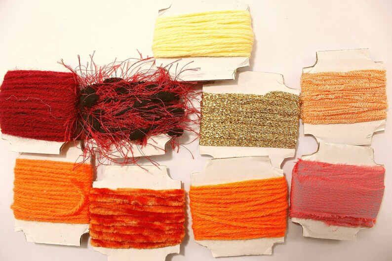 K&ouml;nnte beinhalten: Sortiment an bunten Garnrollen auf wei&szlig;em Karton. Die Garnfarben umfassen Rot, Orange, Gelb, Gold und Rosa.