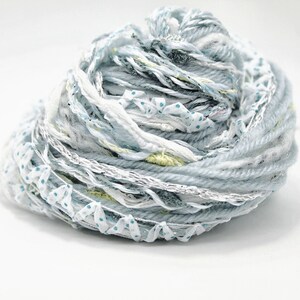 Puede incluir: Ovillo de hilo mixto en tonos azul claro, blanco y plateado. El hilo incluye varias texturas, como hebras gruesas, cintas con lunares azules e hilos metálicos. Ideal para tejer u otras manualidades.