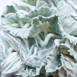 Può includere: Primo piano di un'opera d'arte tessile con varie trame e colori. Il pezzo include tessuto verde chiaro, arricciato con un motivo a diamante, intrecciato con fili bianchi e argentati. L'effetto complessivo è morbido e strutturato.