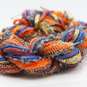 Puede incluir: Primer plano de una madeja de hilo colorida. El hilo presenta hebras naranjas, azules, grises y doradas entrelazadas. El hilo es una mezcla de texturas, incluyendo una malla metálica y una textura difusa.