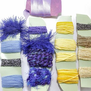 May include: A collection of colorful yarn samples in shades of blue, yellow, and metallic silver and gold. The yarn is arranged on light green cardstock, with a lavender ribbon and fabric strip at the top. The textures vary from smooth to fuzzy.