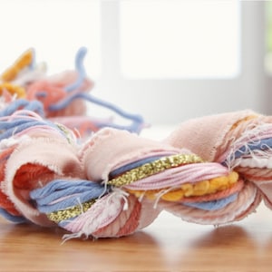 Peut inclure: Gros plan sur un textile tressé aux couleurs pastel. La tresse est composée de tissus et de fils roses, bleus, oranges et dorés. Le textile est posé sur une surface en bois, avec un arrière-plan flou.