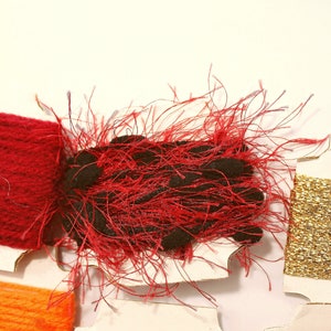 K&ouml;nnte beinhalten: Sortiment von Stickgarnrollen in Rot, Orange, Gold und Schwarz. Die rote Rolle ist ein flauschiger, strukturierter Faden. Die orangefarbene und goldene Rolle sind glatt und gl&auml;nzend.