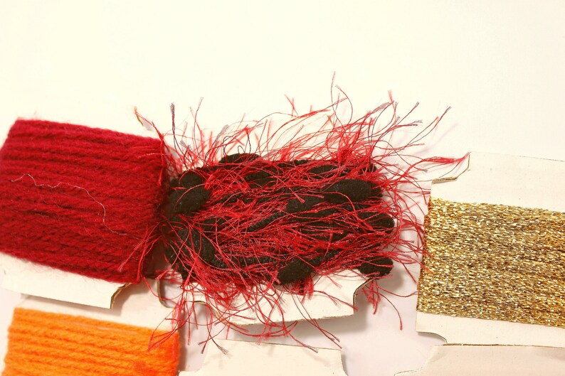 K&ouml;nnte beinhalten: Sortiment von Stickgarnrollen in Rot, Orange, Gold und Schwarz. Die rote Rolle ist ein flauschiger, strukturierter Faden. Die orangefarbene und goldene Rolle sind glatt und gl&auml;nzend.