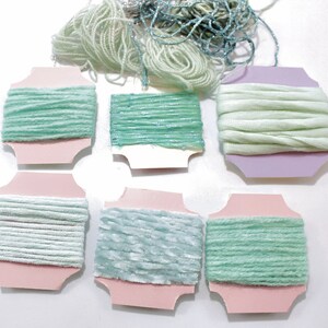 May include: Assortment of seven spools of yarn in various shades of green and white. The yarn is wound around cardboard spools with pink and purple labels.