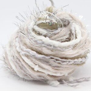 May include: A ball of yarn with a mix of white, beige, and silver threads. The yarn has a fluffy texture and is decorated with silver tinsel.