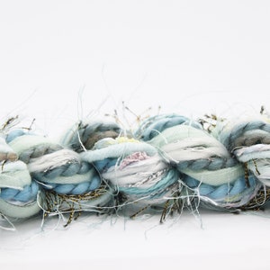 May include: Close-up of a thick, textured yarn in shades of blue, green, and white. The yarn has a braided appearance with strands of silver and gold metallic thread. The overall effect is soft and luxurious, suitable for crafting projects.