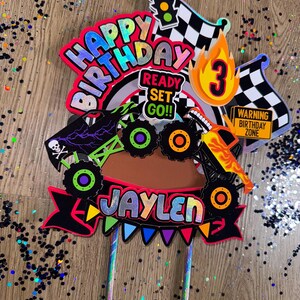 May include: A colorful cake topper featuring a monster truck with flames, a checkered flag, a traffic light, and the words "Happy Birthday", "Ready Set Go!" and "Jaylen". The topper also includes the number "3" and a warning sign that says "Warning Birthday Zone".