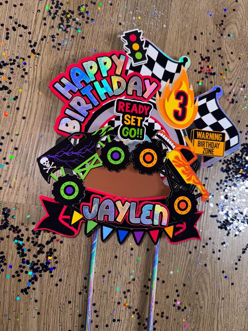 May include: A colorful cake topper featuring a monster truck with flames, a checkered flag, a traffic light, and the words "Happy Birthday", "Ready Set Go!" and "Jaylen". The topper also includes the number "3" and a warning sign that says "Warning Birthday Zone".