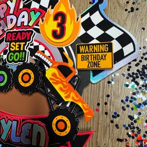 May include: A monster truck cake topper with a yellow and orange monster truck, a checkered flag, a fire with the number 3, a sign that says "Warning Birthday Zone", and a banner that says "Ready Set Go!"