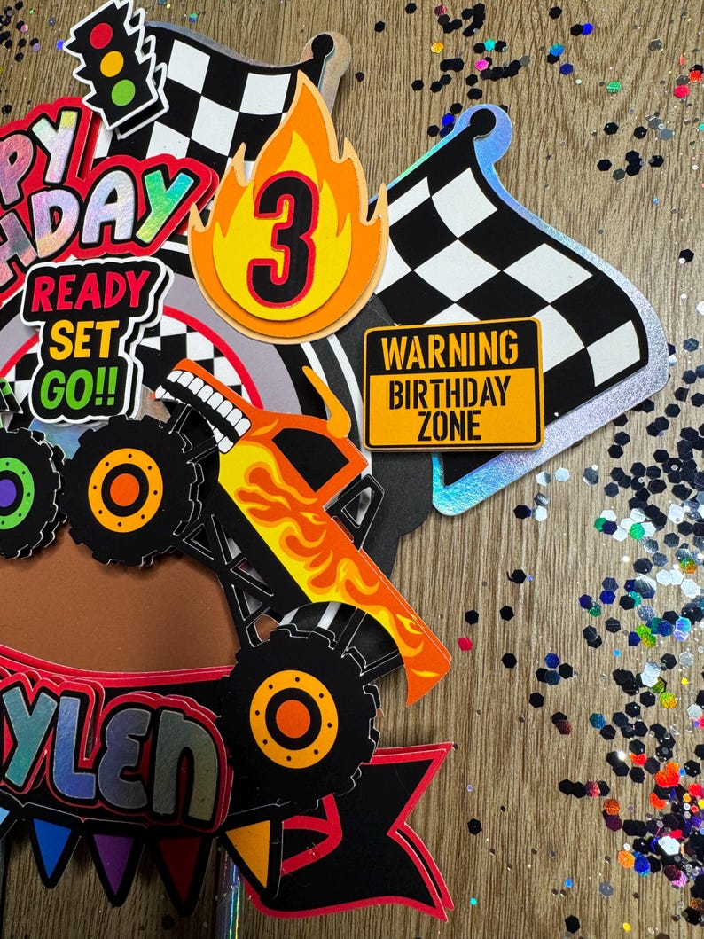 May include: A monster truck cake topper with a yellow and orange monster truck, a checkered flag, a fire with the number 3, a sign that says "Warning Birthday Zone", and a banner that says "Ready Set Go!"