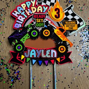 May include: A colorful monster truck themed cake topper with the words "Happy Birthday", "Ready Set Go!" and "Jaylen". The topper also includes a checkered flag, a traffic light, a number 3, and a sign that says "Warning Birthday Zone".