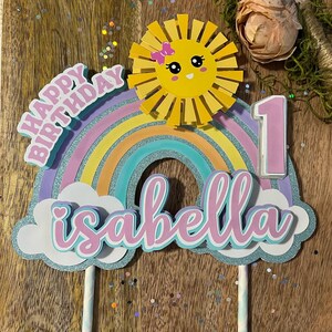 Może przedstawiać: Topper na tort w kształcie tęczy z uśmiechniętym słońcem, r&oacute;żową jedynką i imieniem "Isabella" w r&oacute;żowych literach. Topper znajduje się na drewnianym tle z brokatem.