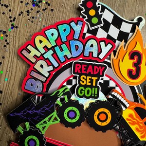 May include: Monster truck themed cake topper set with a red, blue, and green "Happy Birthday" banner, a checkered flag, a traffic light, a monster truck silhouette, and a number 3 with flames. The set also includes a "Ready Set Go!" banner.