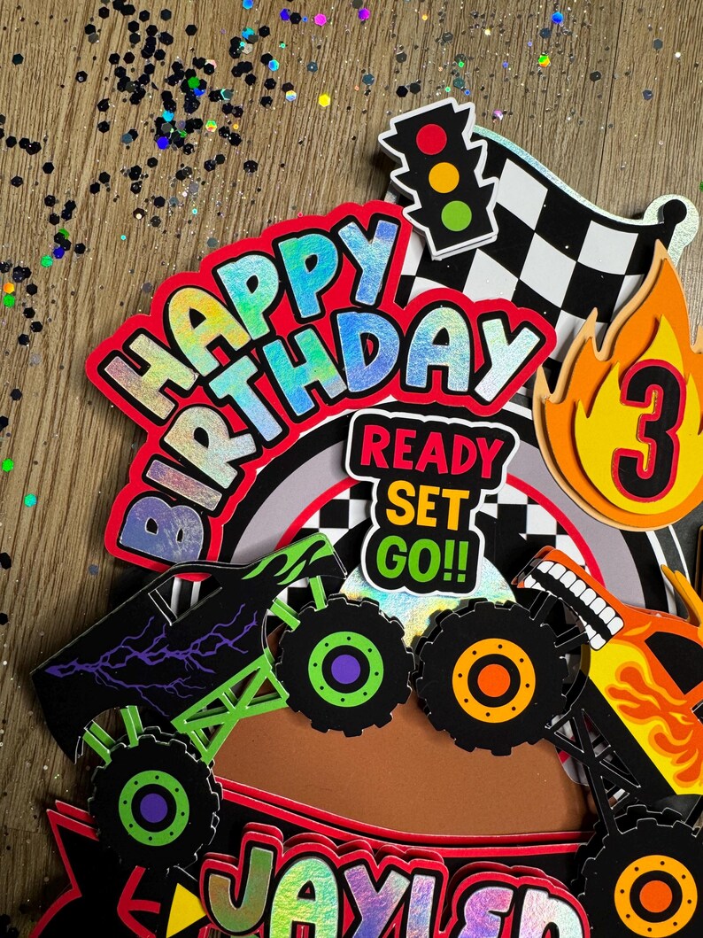 May include: Monster truck themed cake topper set with a red, blue, and green "Happy Birthday" banner, a checkered flag, a traffic light, a monster truck silhouette, and a number 3 with flames. The set also includes a "Ready Set Go!" banner.