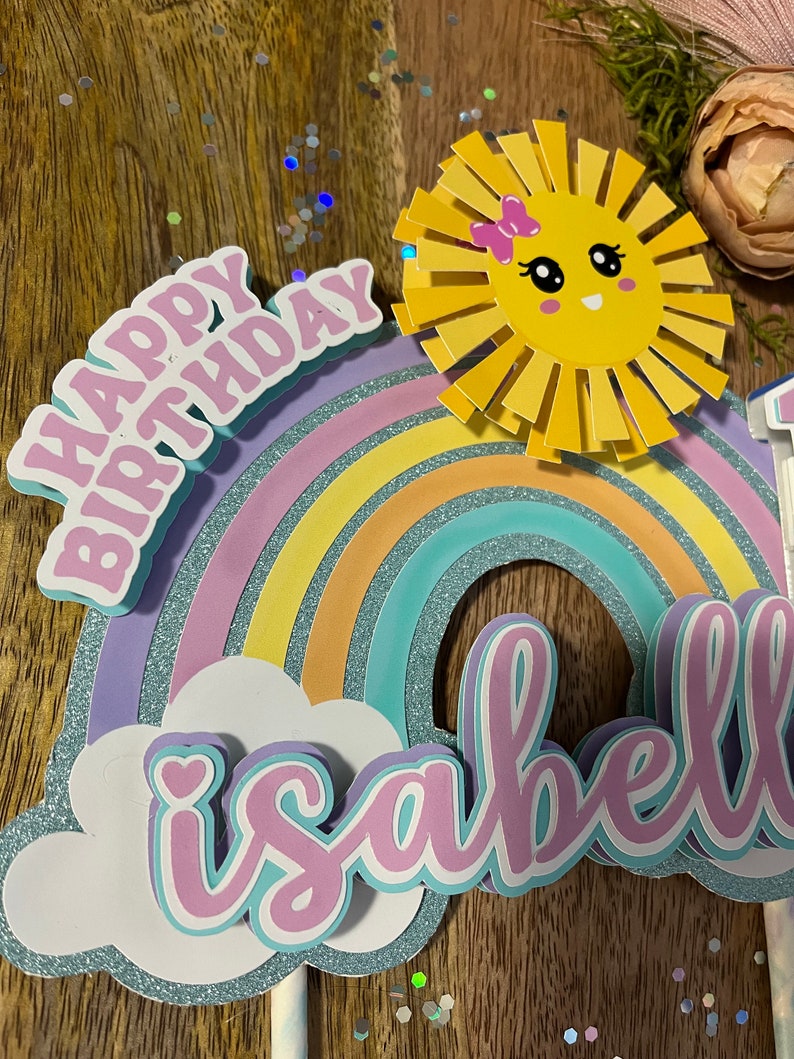 Może przedstawiać: Topper na tort w kształcie tęczy z słońcem, chmurą i imieniem "Isabella" w r&oacute;żowych literach. Tęcza jest wykonana z pastelowych kolor&oacute;w i ma błyszczące elementy. Słońce ma r&oacute;żową kokardkę i uśmiechniętą buźkę. Chmura ma małe r&oacute;żowe serduszko. Topper na tort jest idealny na przyjęcie urodzinowe.