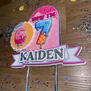 May include: A colorful birthday cake topper with the name "KAIDEN" in blue glitter. The topper features the numbers "6" and "7" designed as ice cream cones, with the text "NOW I'M". The design is pink, blue, and orange.